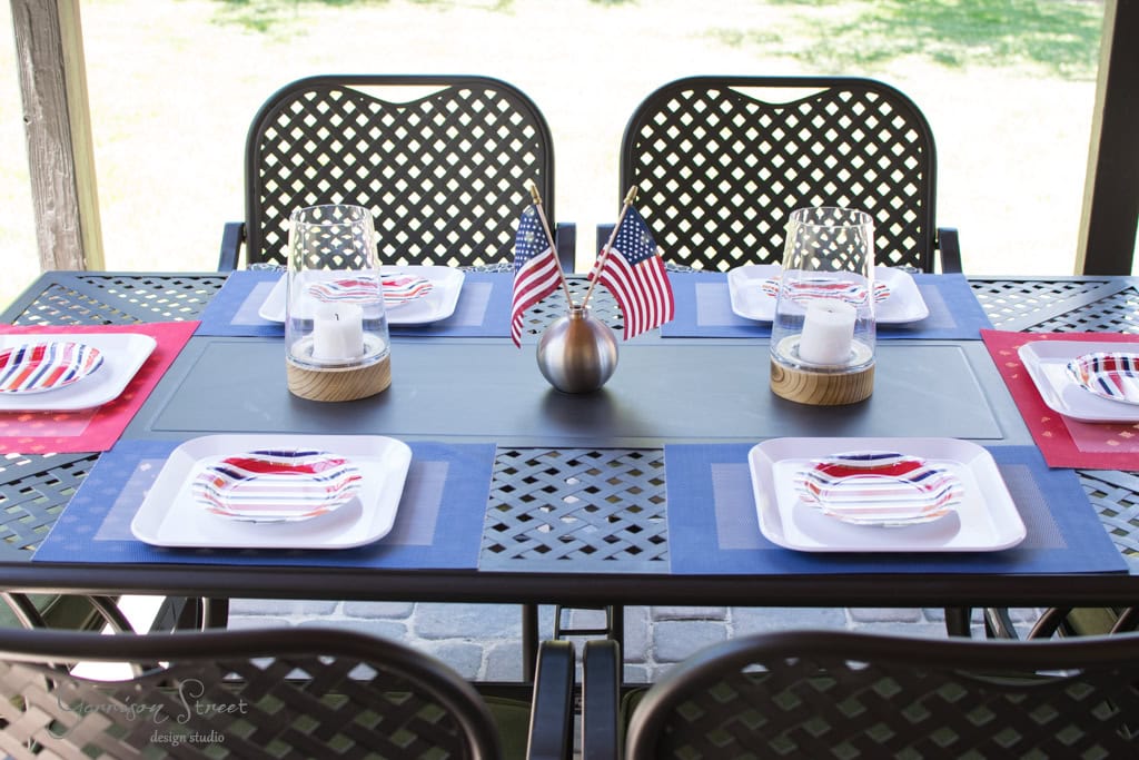 A patio table set for a meal with four chairs. It features red, white, and blue themed placemats, plates, and miniature American flags. The centerpieces include glass lanterns with candles. The setting suggests a celebration or gathering.