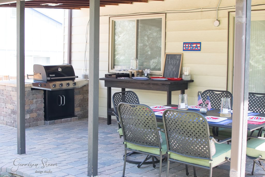 Outdoor patio with a grill on a brick platform, a long table set with American flag-themed placemats, and a small chalkboard sign on a wooden counter. The patio is covered with a wooden pergola.