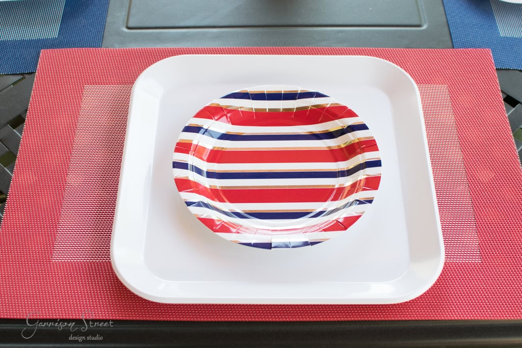 A table setting features a square white plate with a smaller round paper plate on top. The paper plate has red, white, and blue stripes. The placemat is red, with a textured rectangular design, and it all rests on a dark surface.