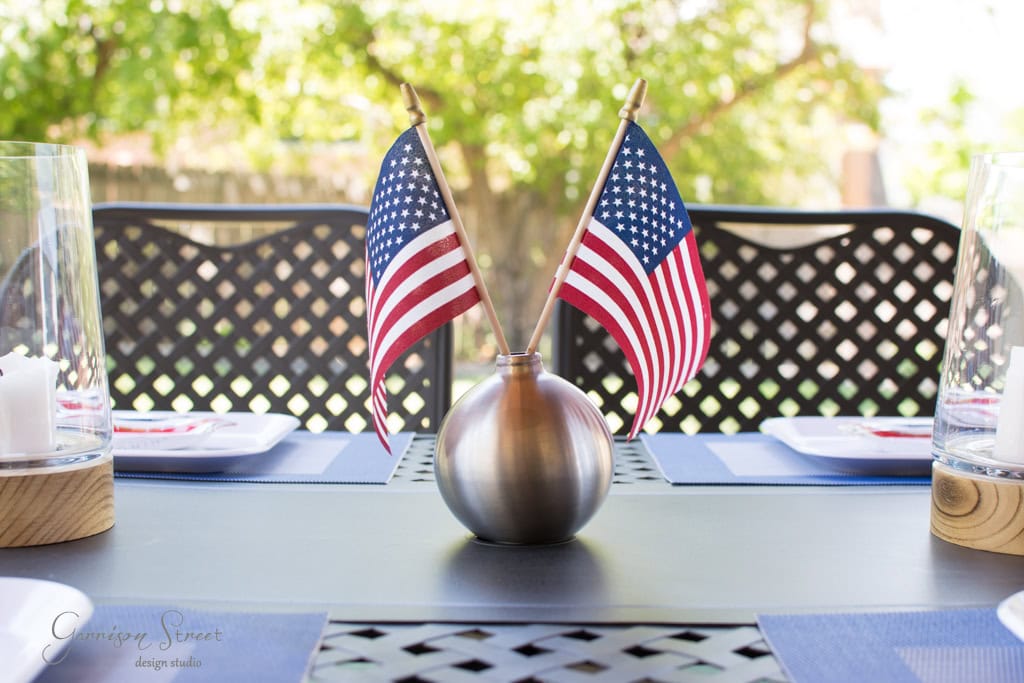 A table set outdoors with two small American flags in a round vase as the centerpiece. Plates and glasses are arranged neatly, and sunlight filters through the trees, creating a bright and festive atmosphere.