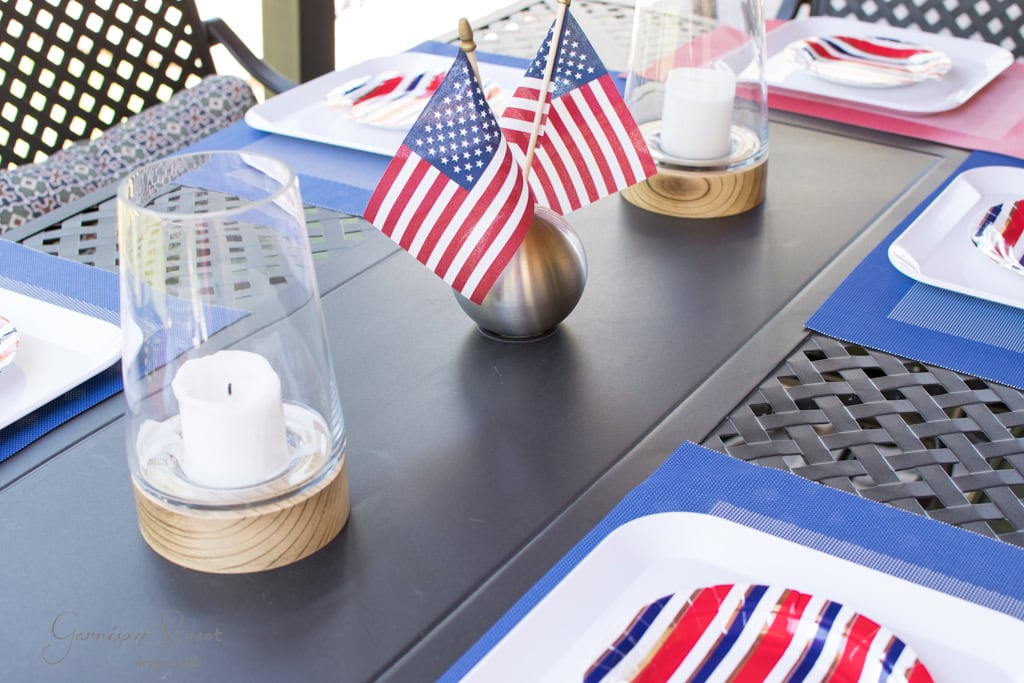 A table set for a patriotic celebration features American flag-themed plates, small American flags in the center, and glass candle holders with white candles. Blue placemats add a pop of color on the black table.