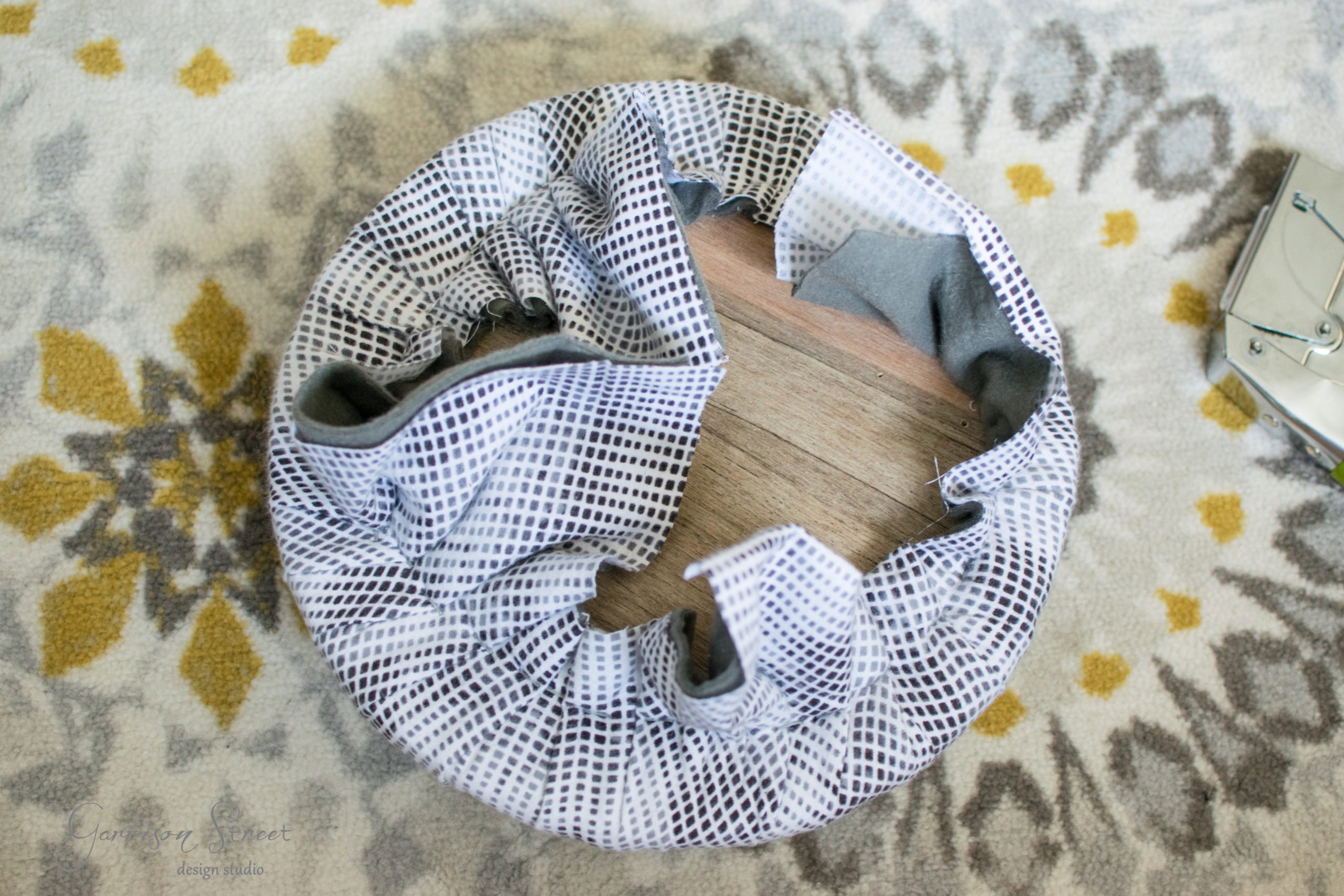 A crumpled, white mesh laundry basket with a gray interior sits on a wooden floor, partially surrounded by a patterned rug with yellow and gray designs. A metal object is visible in the corner.