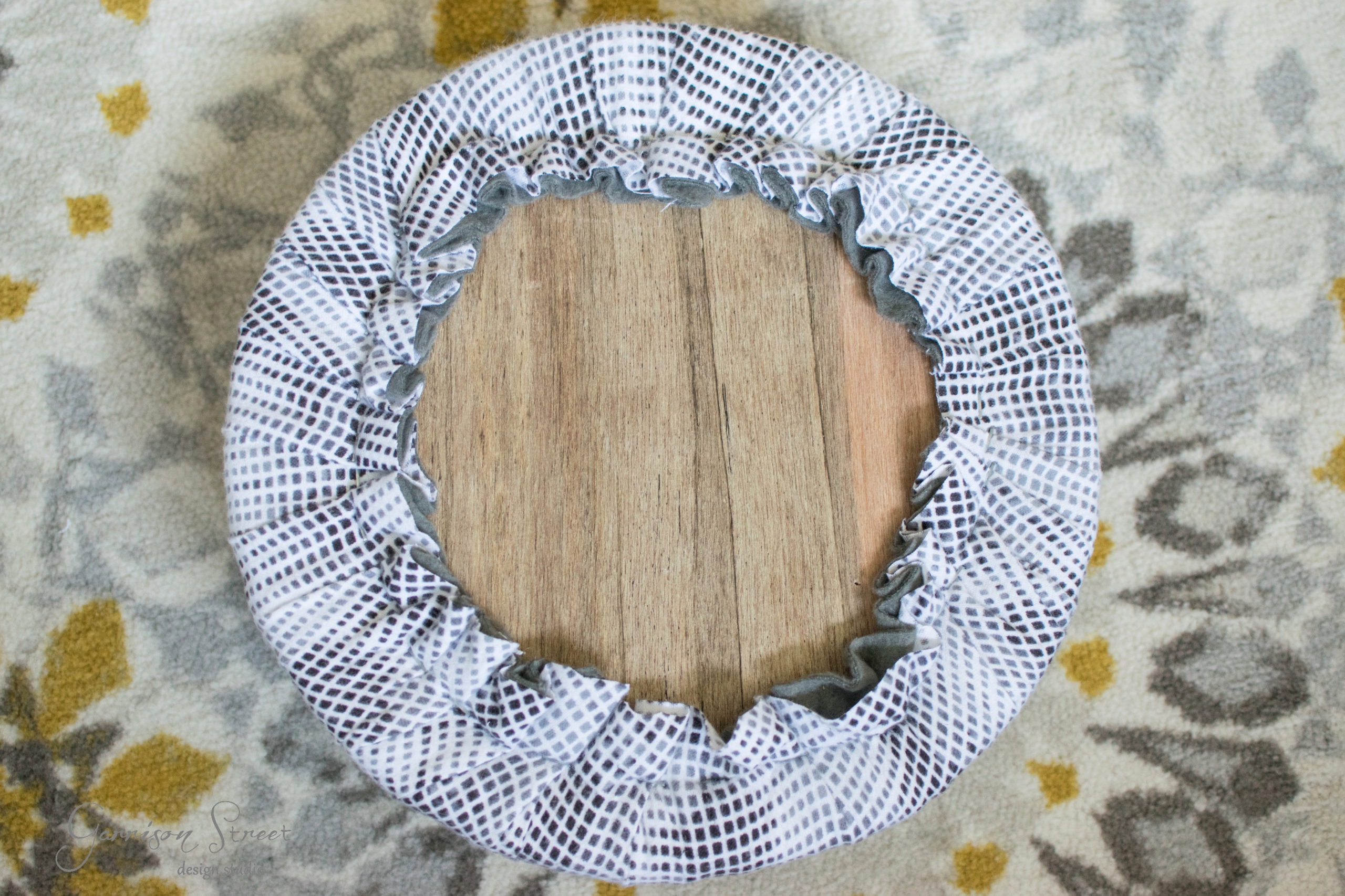 A round, wood-textured stool seat with a ruffled cover featuring a white and black dot pattern is centered on a floral-patterned carpet with gray and yellow tones.