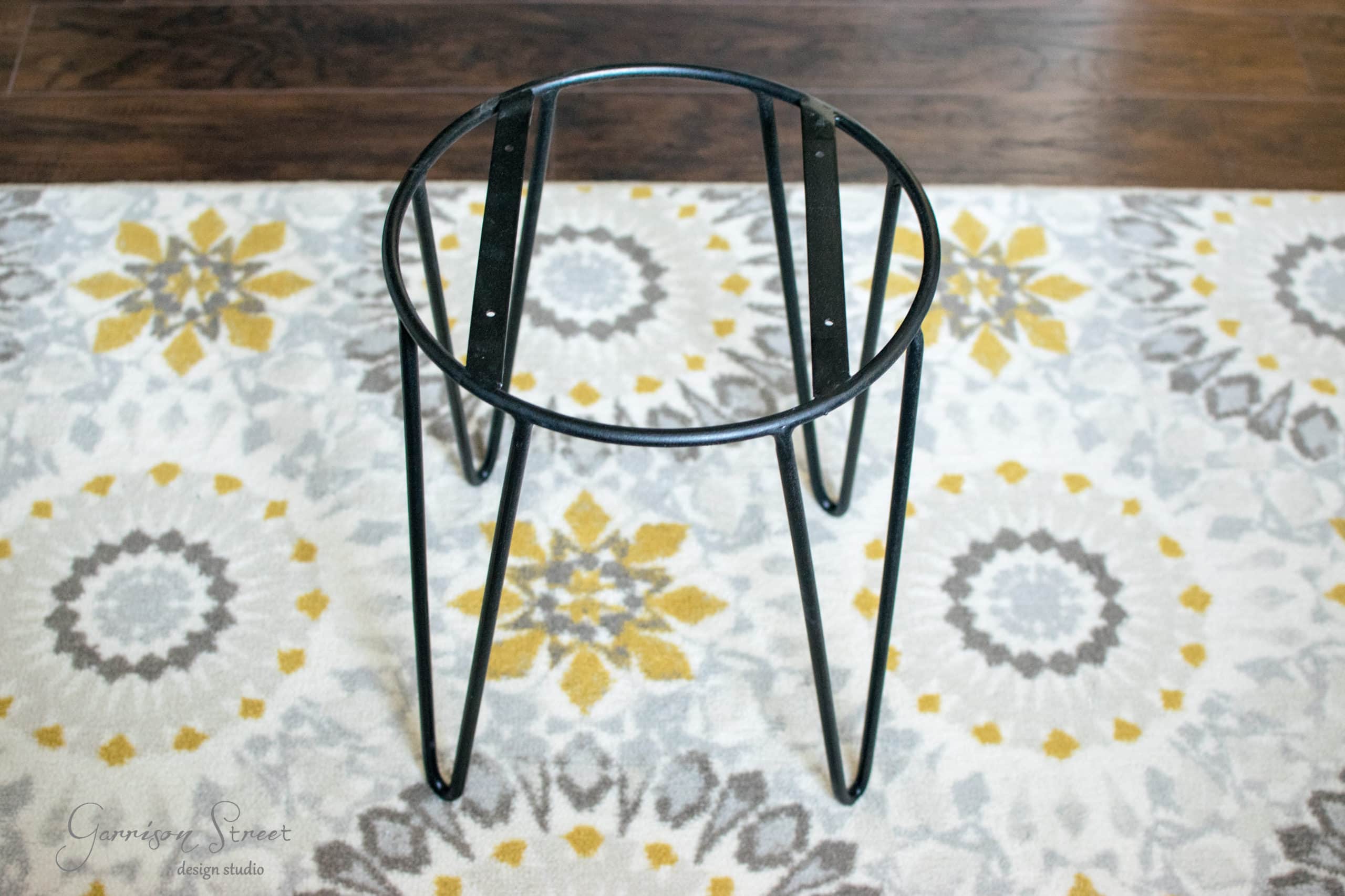 A round, black metal stool frame with hairpin legs placed on a patterned rug featuring yellow, gray, and white floral designs. The floor beneath is dark wood.