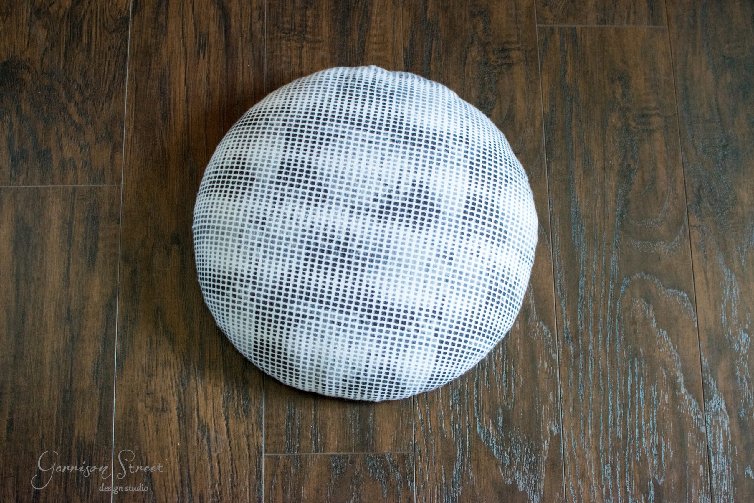 A round cushion with a black and white grid pattern sits on a dark wooden floor.