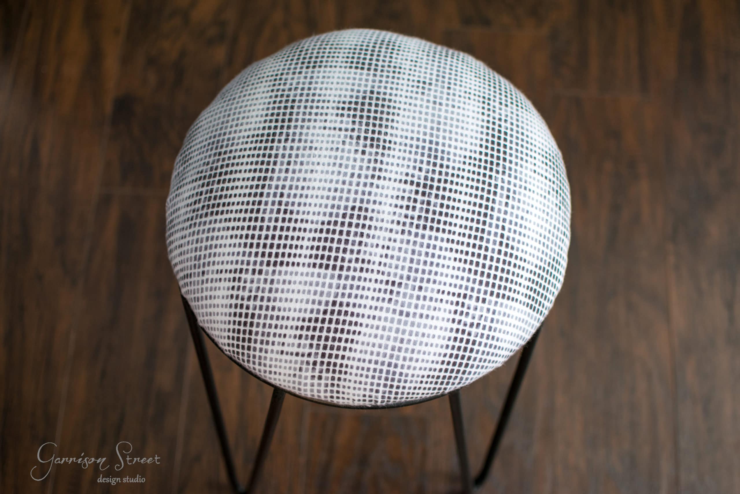 A round, modern stool with a metal frame and a checkered pattern cushion stands on a wooden floor, viewed from above.