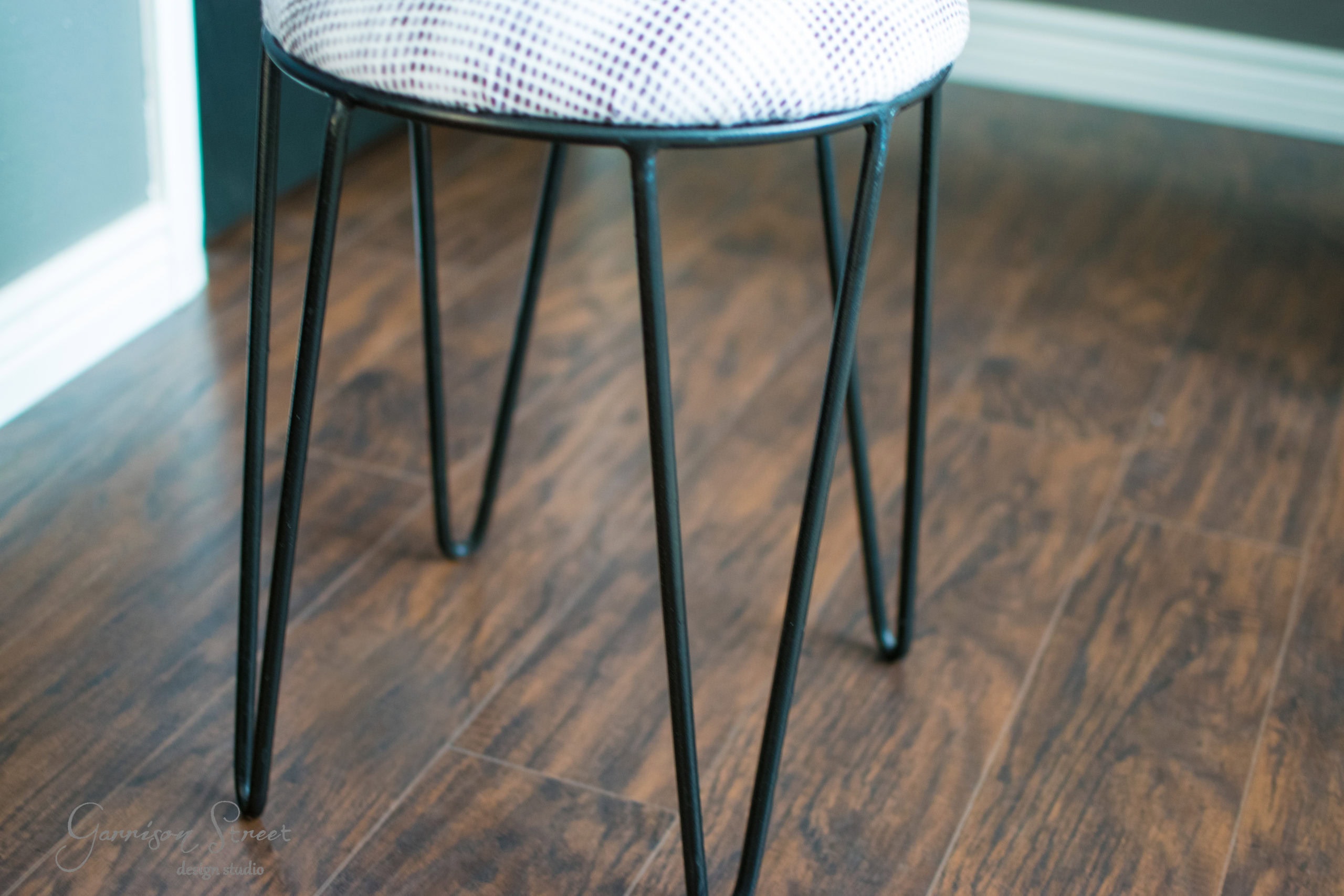 A modern stool with a patterned cushion top and black metal legs is placed on a wooden floor. The legs are slim and angled, giving a minimalist, contemporary design.