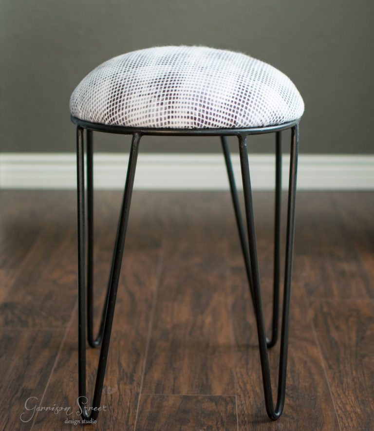 A small stool with a round, white-snake-patterned seat and black metal hairpin legs, placed on a wooden floor against a gray wall.