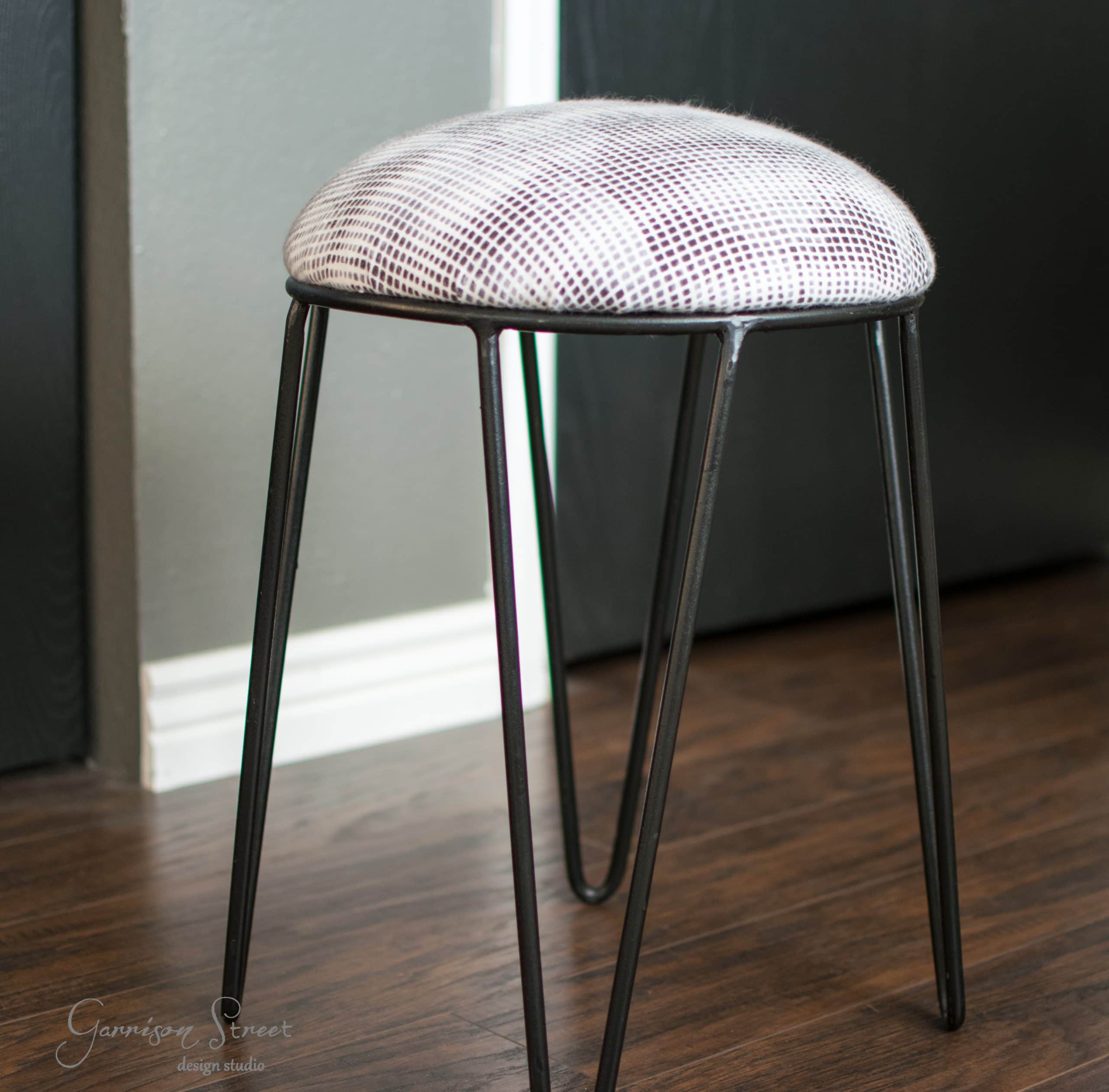 A modern stool with a round, checkered fabric seat and sleek black metal legs sits on a wooden floor against a gray wall.