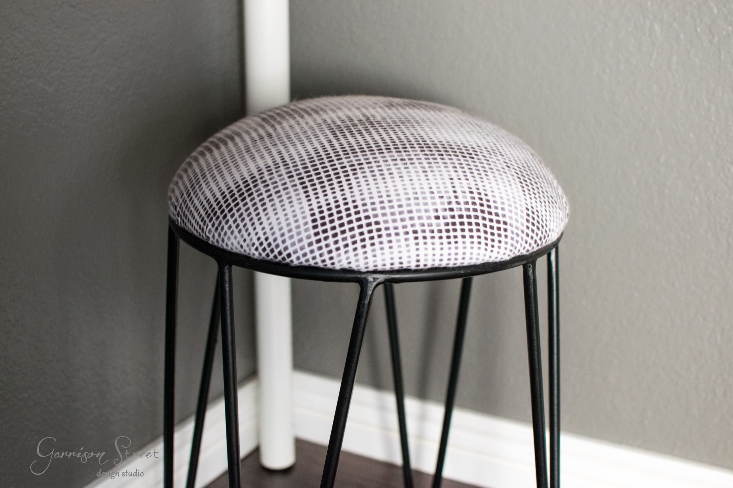 A round stool with a textured gray cushion and black metal legs stands in front of a gray wall. The stool is placed on a dark wooden floor near a white baseboard.