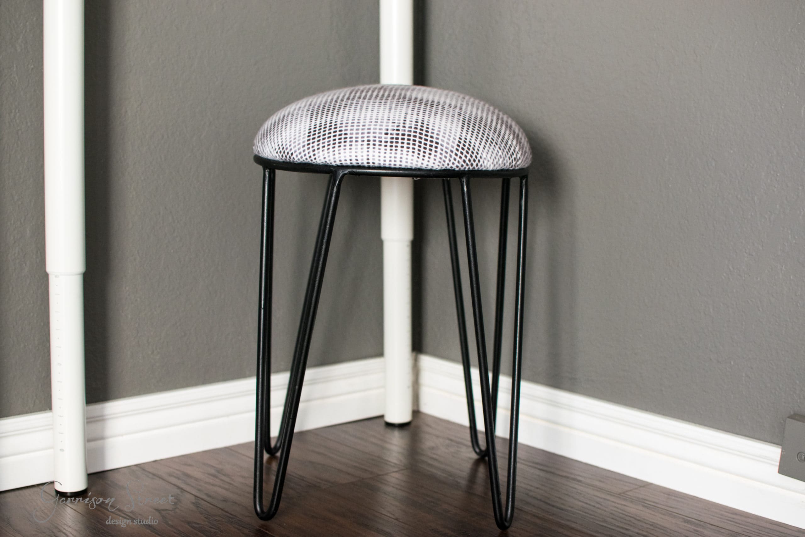 A small stool with a round, upholstered seat featuring a black and white pattern. It has three black metal hairpin legs and is positioned in the corner of a room with wooden flooring and gray walls.