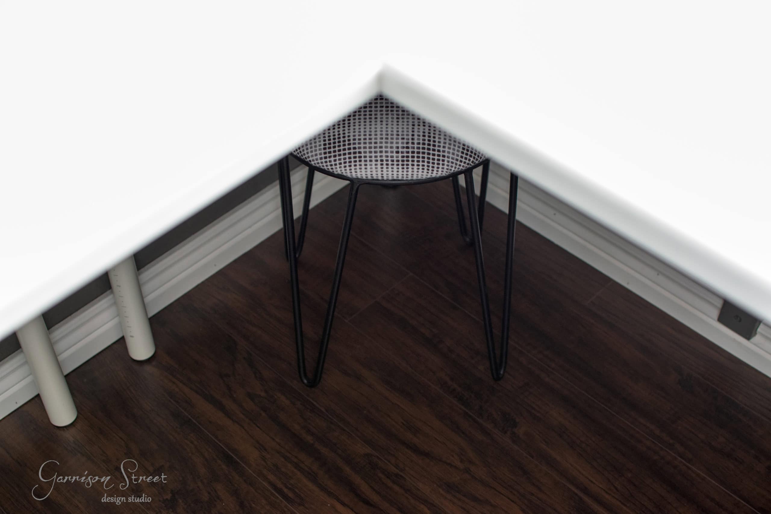 A small metal stool with a perforated seat is tucked under a white corner desk. The floor is dark wood, and part of the white wall baseboard is visible. The stool has thin black legs.