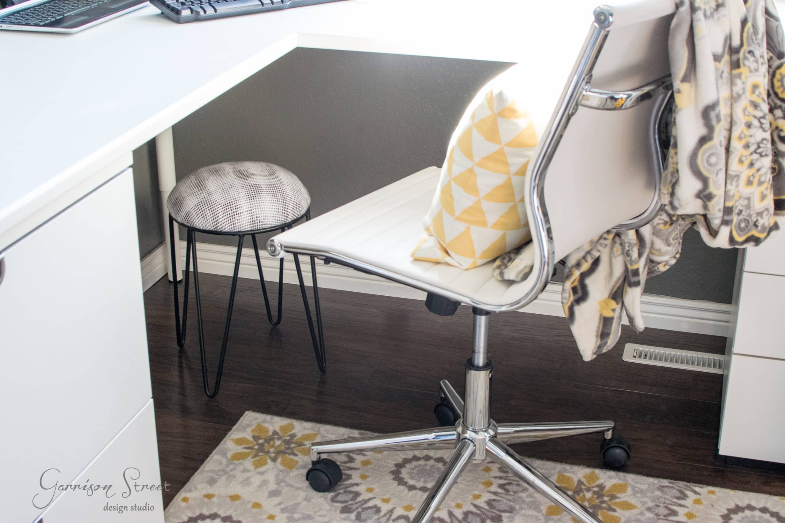 A home office setup with a white desk and a white swivel chair. There's a yellow patterned cushion on the chair and a patterned throw draped over the back. A small stool with a round cushion is tucked under the desk. A floral rug covers the dark floor.