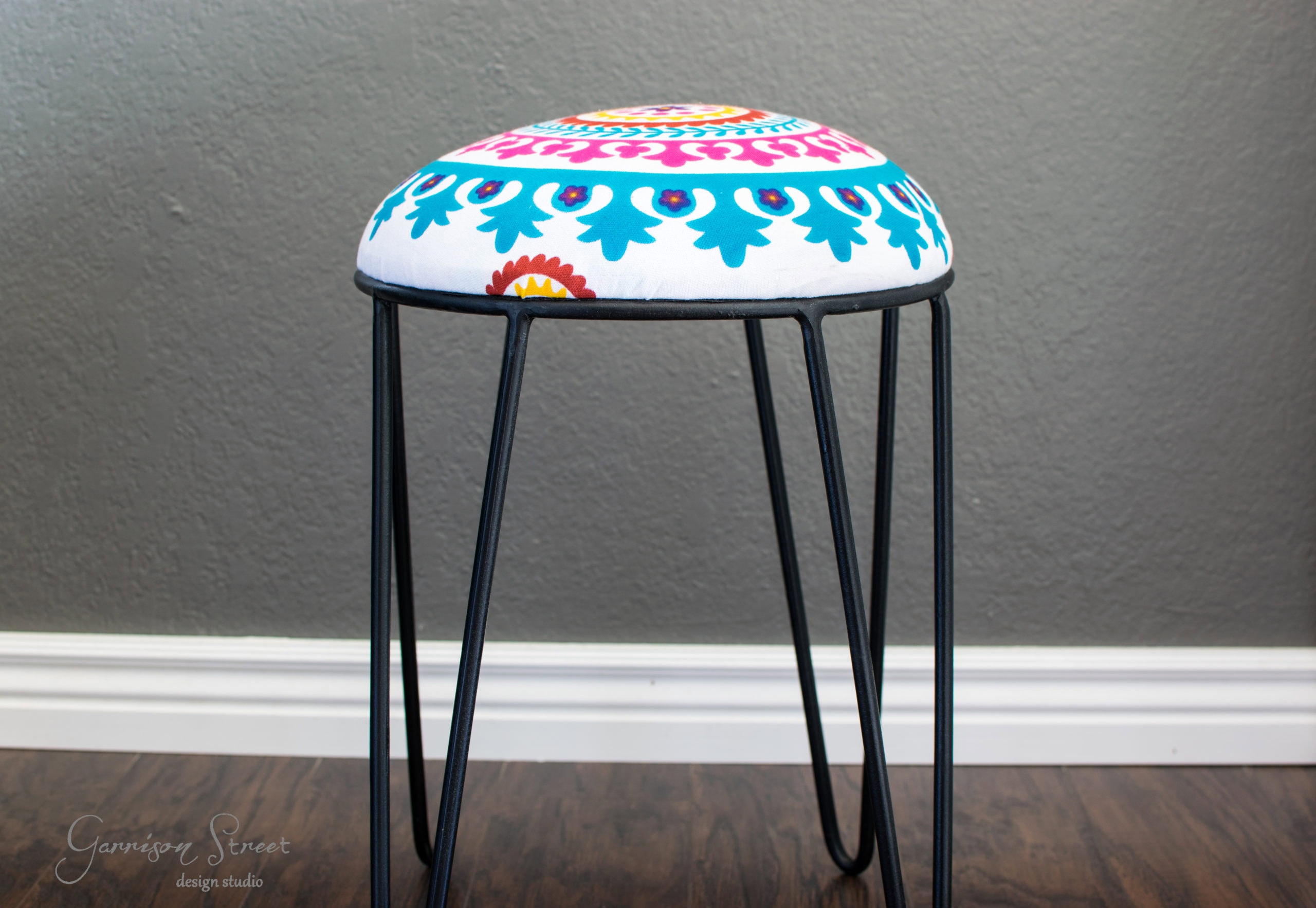 A small stool with thin black metal legs and a round, colorful cushion featuring blue, pink, and orange patterns on a white background, placed on a wooden floor against a gray wall.