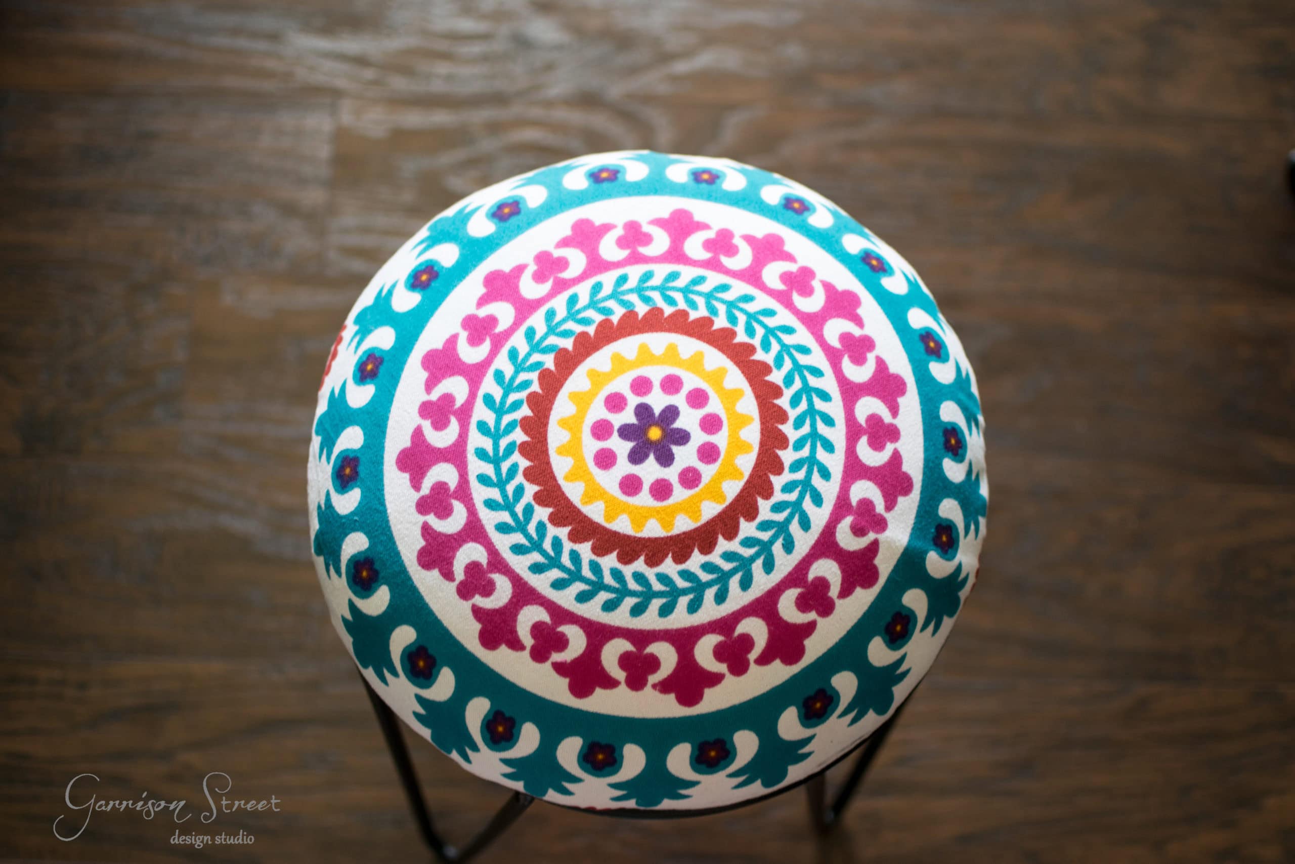 Top view of a colorful round stool with a vibrant mandala pattern featuring blue, pink, and orange designs on a wooden floor.