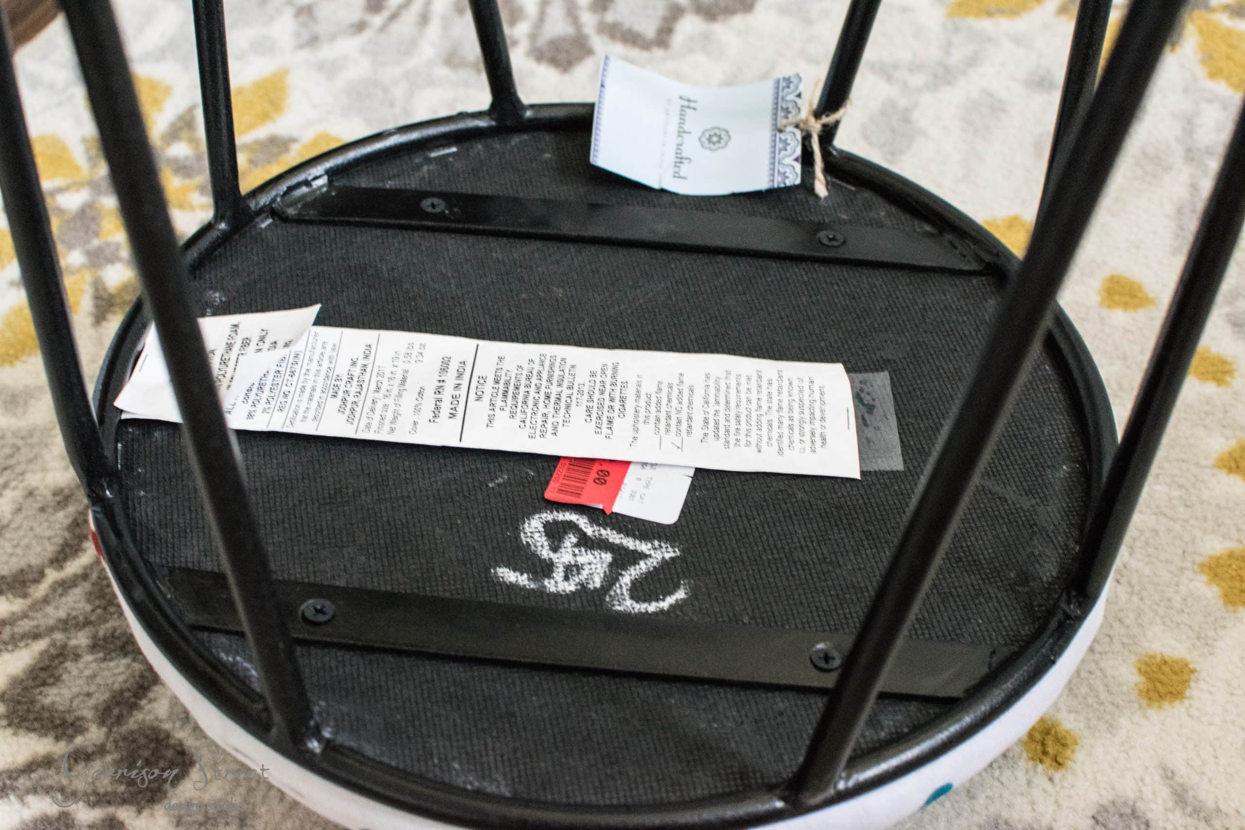 The image shows the underside of a round stool or small table with black metal legs. Several tags and labels with text are attached, along with a chalk or paint marking. It's placed on a patterned rug with yellow and gray designs.