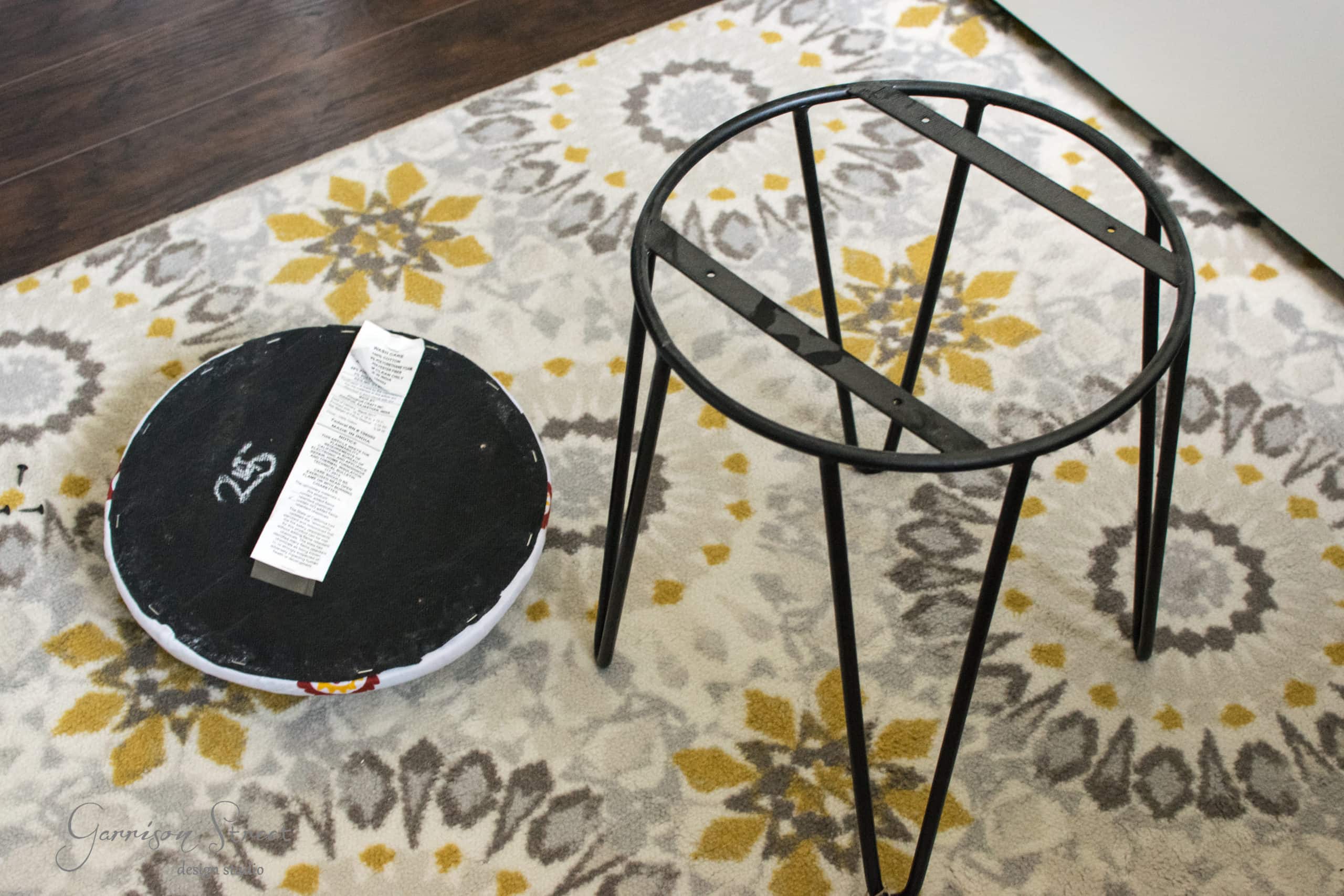 A black metal stool frame lies on a patterned carpet next to a circular stool seat with an attached instruction sheet.