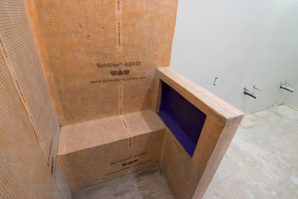 A partially constructed bathroom shower features Schluter-KERDI waterproofing membrane on the walls, with a built-in niche and a shower bench. Unfinished flooring and exposed plumbing are visible.
