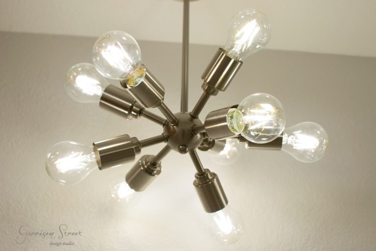 A modern chandelier with multiple exposed bulbs extends from a central metallic hub, resembling a starburst, against a plain ceiling. The bulbs are lit, casting a warm glow.