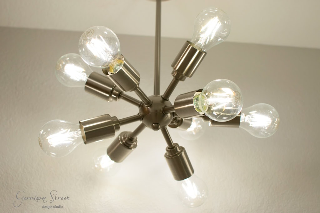 A modern chandelier with multiple exposed bulbs extends from a central metallic hub, resembling a starburst, against a plain ceiling. The bulbs are lit, casting a warm glow.