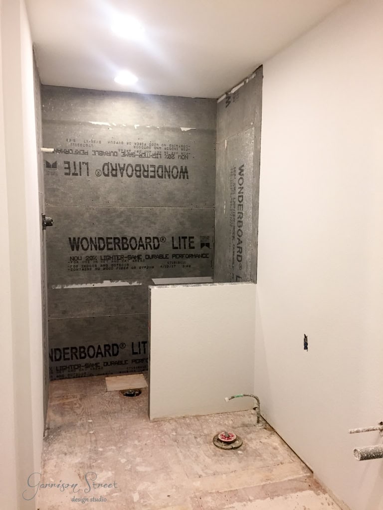 Bathroom under construction with Wonderboard Lite panels on the walls. The floor is unfinished, with visible plumbing and no fixtures. Recessed ceiling lights provide illumination.