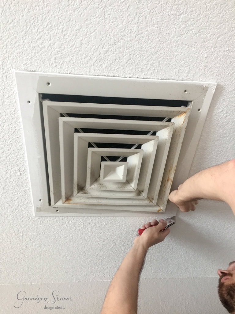 A person uses a screwdriver to unscrew a rectangular white ceiling air vent for cleaning. Rust stains are visible on the vent.