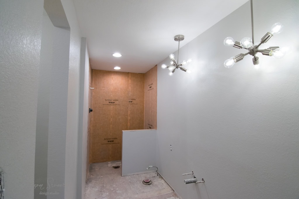 A partially completed bathroom with bare walls and floors. There are exposed plumbing fixtures and two modern chandeliers. The room has recessed lighting and an unfinished walk-in shower area with brown wallboard.