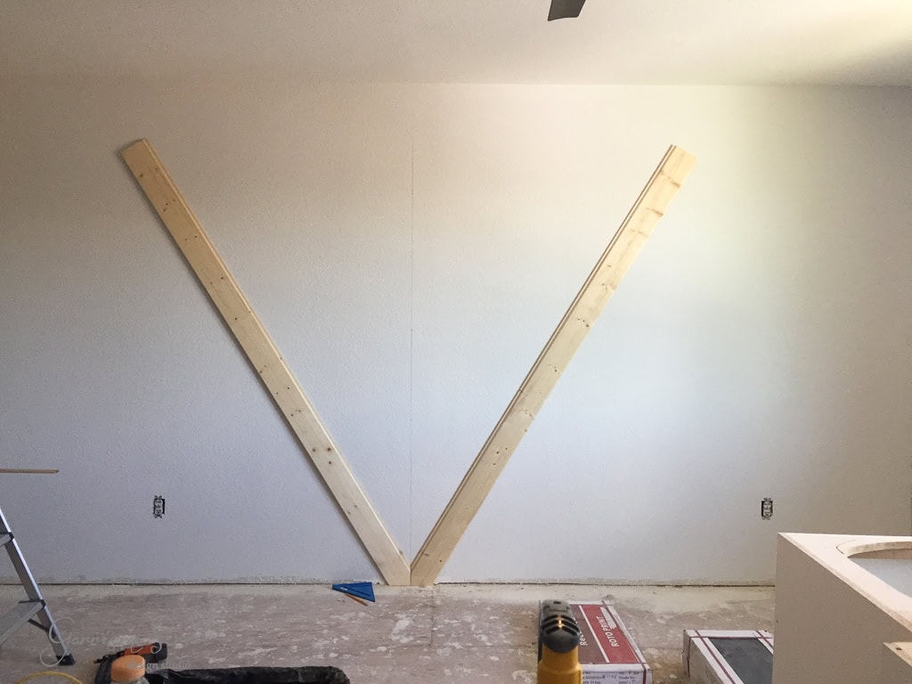 Two wooden beams arranged in a large V shape are mounted on a white wall. The wall has unpainted patches and the floor is bare. Construction tools and materials are scattered at the bottom of the image.