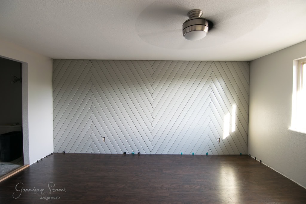 A minimalist room with a herringbone-patterned accent wall painted light gray. The floor is dark wood, and there's a modern ceiling light. Sunlight enters through a window on the right, casting soft shadows.