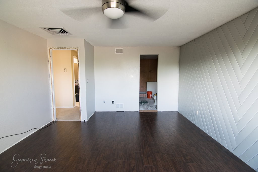 A partially renovated room with dark wood flooring and a herringbone accent wall. An open doorway reveals a small area with remodeling tools, including a red bucket. A ceiling fan is mounted overhead.