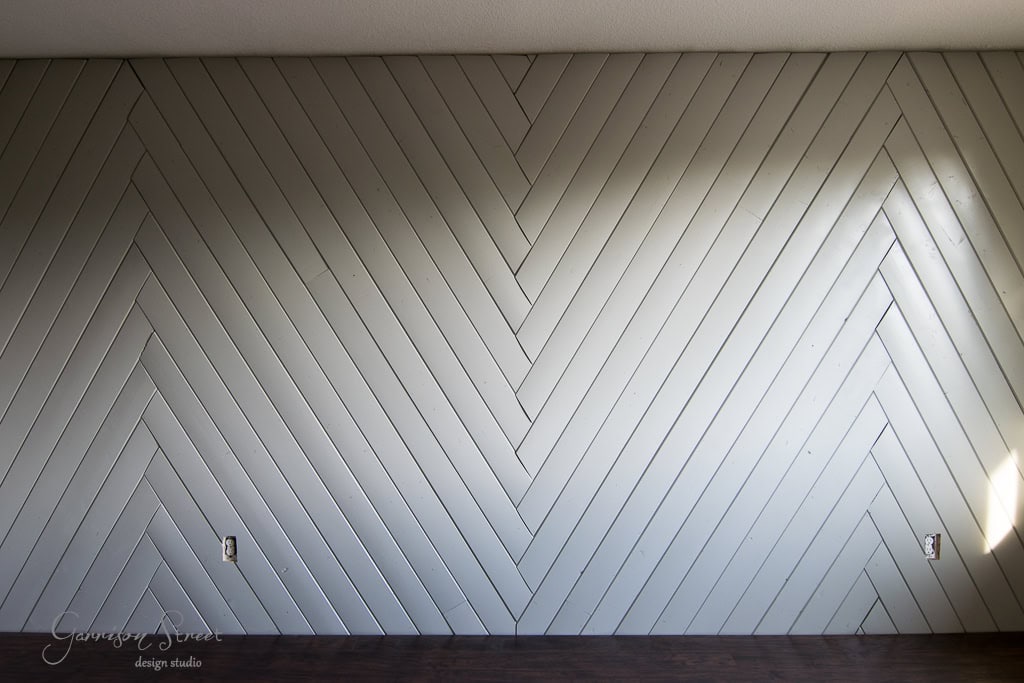 A wall with a light gray herringbone wood pattern, featuring two empty electrical outlets at the bottom. The floor is dark wood, and sunlight casts a diagonal shadow across the wall.