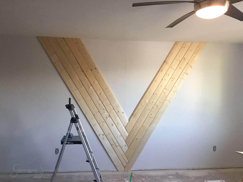 A partially completed wall design with wooden planks forming a large "V" shape on a plain white wall. A step ladder is positioned to the left, and a ceiling fan with a light is visible above. The floor appears unfinished.
