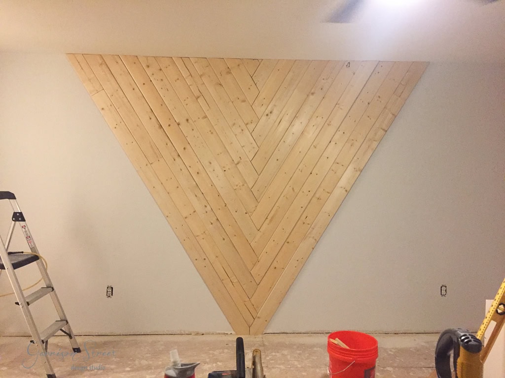 A wall with a large wooden chevron pattern made of light-colored wood planks. A ladder and a red bucket are on the unfinished floor. The room appears to be undergoing renovation.