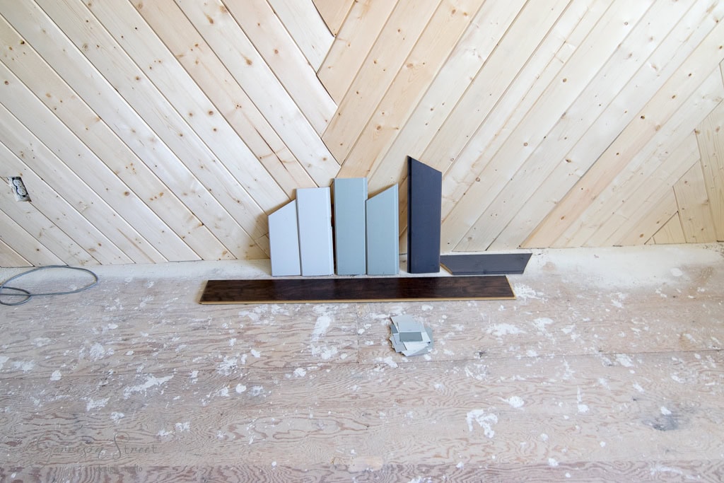 A herringbone wood wall with various paint swatches leaning against it. The swatches are in shades of gray, blue, and black. A dark wooden floorboard and a small gray paint sample rest on a dusty plywood floor.
