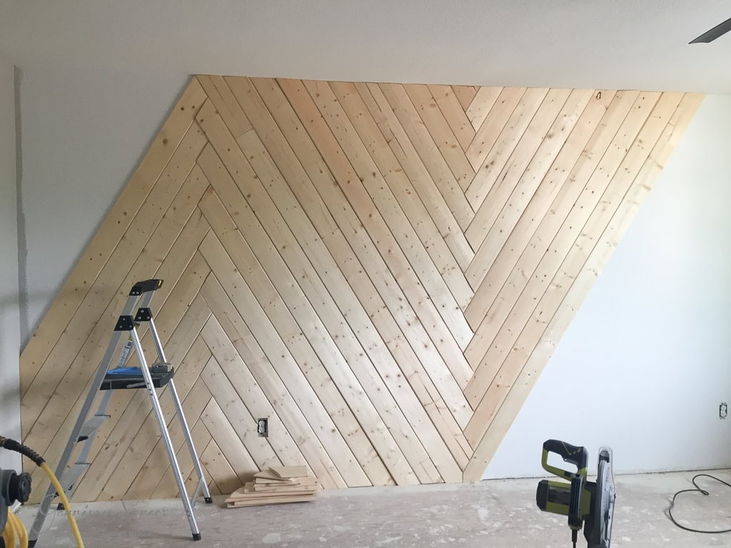 A modern wood accent wall under construction with a chevron pattern made from light wood planks. A step ladder and a saw are placed on the unfinished floor, indicating ongoing work.