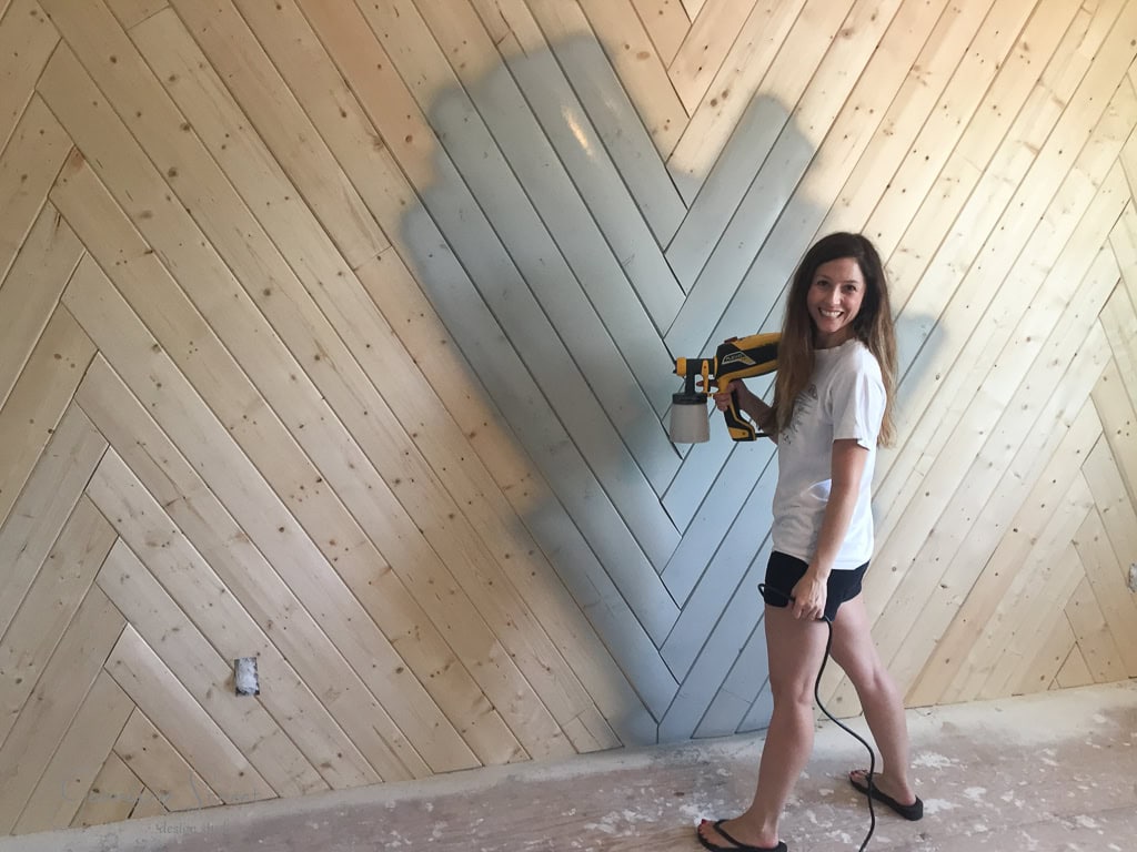 A woman smiles while spray painting a herringbone wooden wall with grayish-blue paint. She holds a paint sprayer and stands on a partially finished floor, wearing a white t-shirt and shorts. The wall features a diagonal plank design.