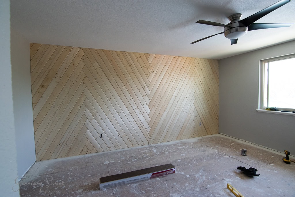 A room with a newly installed wooden herringbone accent wall, an unfinished floor, and a ceiling fan. Tools and materials, like a level and tape measure, are scattered on the floor. Natural light comes in through a window on the right.