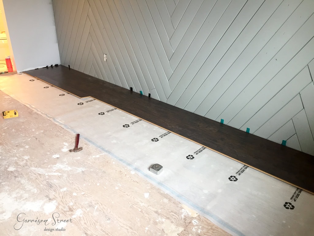 A room with a partially installed dark wood floor. The wall features a chevron pattern in light gray. Tools and materials are visible, including a measuring tape and a red leveling tool on the subfloor.