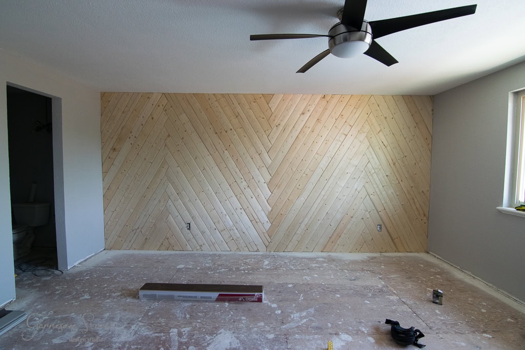 A room with a newly installed herringbone-patterned wood accent wall. The floor is bare, and construction materials are scattered around. A modern ceiling fan is mounted above, and there's an open door to an adjacent room.