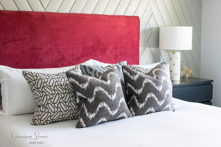 A modern bedroom with a velvet red headboard, patterned gray and white pillows, and a white bedspread. A black nightstand with a white lamp and a decorative gold star are on the side. The wall has a chevron pattern.
