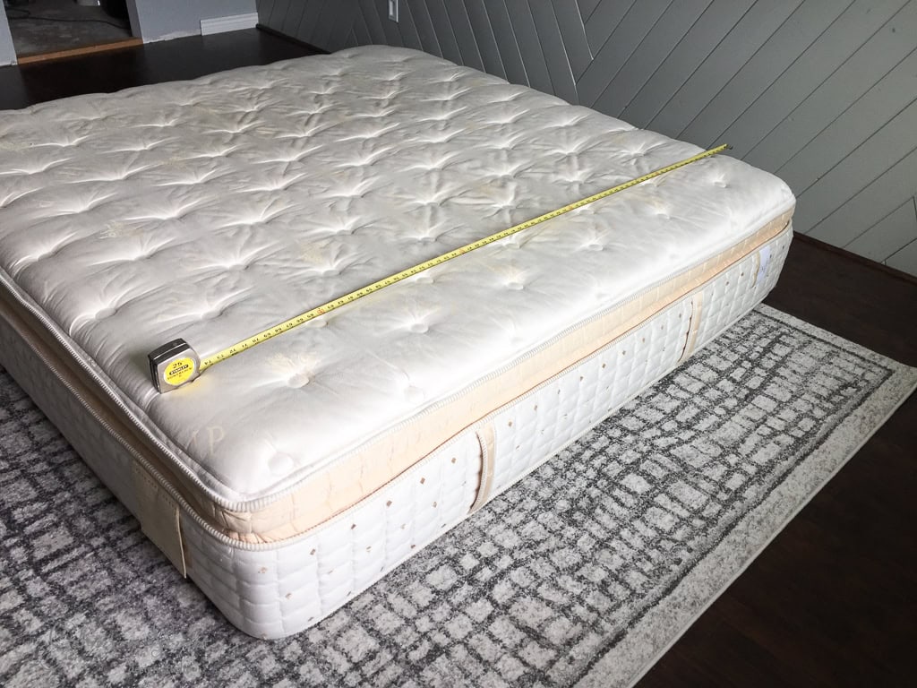 A measuring tape is stretched across a large, plush mattress. The mattress is placed on a decorative area rug with a geometric pattern. The setting appears to be a simple room with wooden flooring and a gray, textured wall.