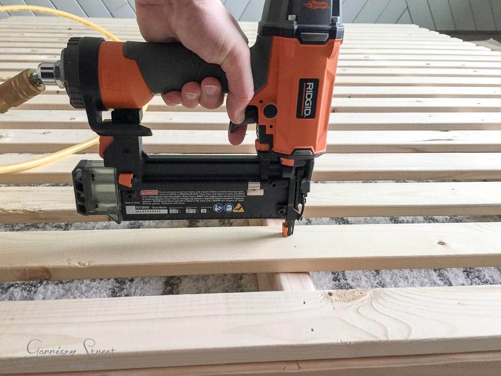 A person uses an orange and black nail gun to attach wooden slats to one another. The slats are laid out on a surface, with the nail gun connected to an air hose.