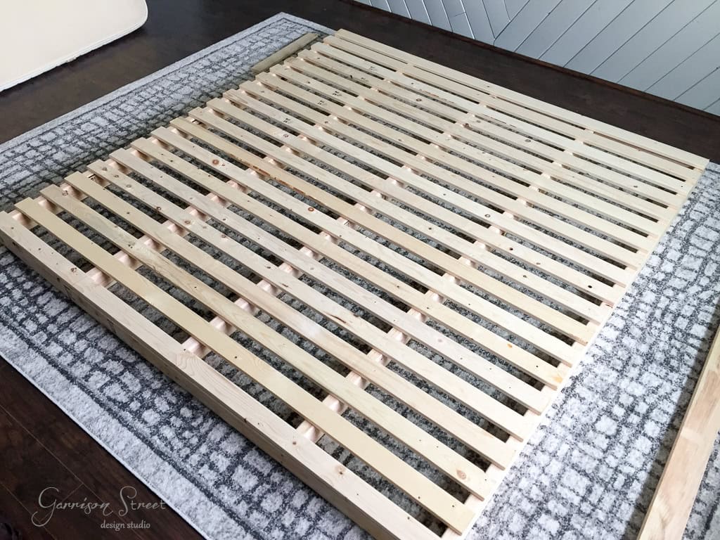 Wooden slatted bed frame laid on a patterned rug in a room. The frame is rectangular with evenly spaced pine slats. The surrounding area features dark wooden flooring and a portion of a light-colored wall in the background.