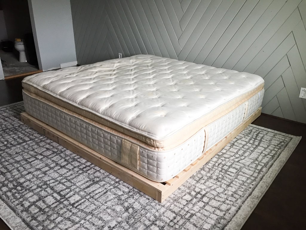 A plush, white mattress on a minimalist wooden bed frame sits atop a textured gray and white rug. The room features a chevron-patterned accent wall and a partially visible doorway in the background.