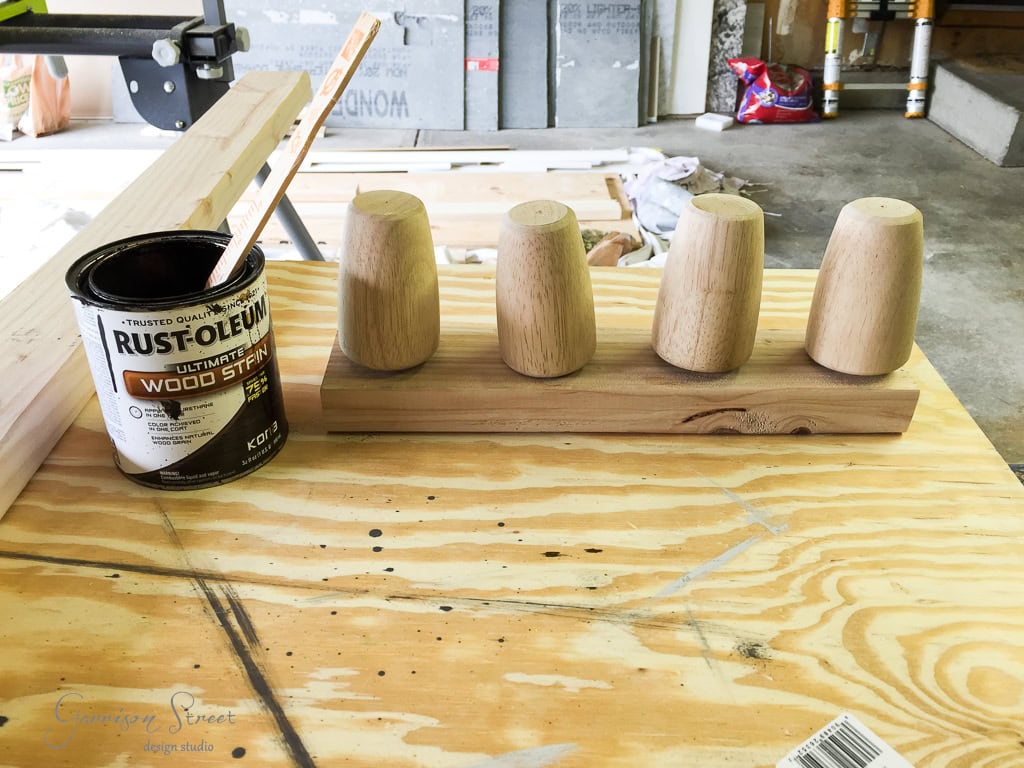 A can of wood stain with a brush lies on a wooden table next to four unfinished wooden legs standing upright on a wooden plank in a workshop.