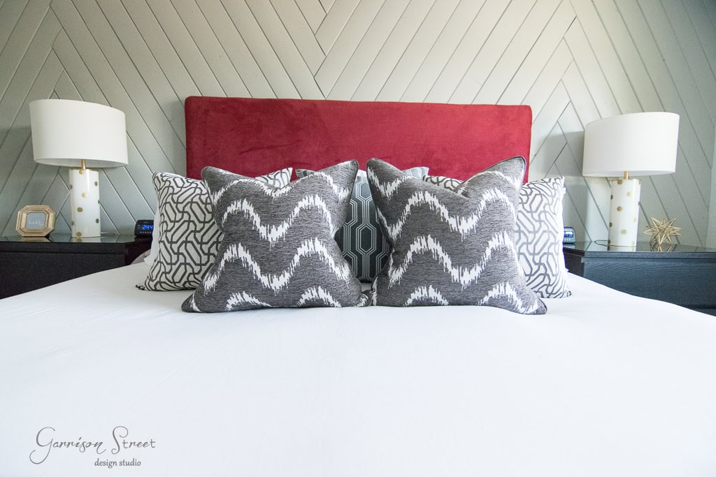 A neatly made bed with a red headboard, featuring patterned gray and white pillows. Two white lamps with gold accents are on dark nightstands on either side. The wall behind has a chevron design.