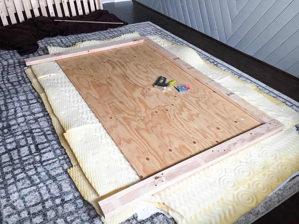 A wooden board lies on a padded surface with two wooden beams placed on top. Various tools and small items, such as a tape measure and sandpaper, are scattered on the board. The setting appears to be a work area or room under renovation.