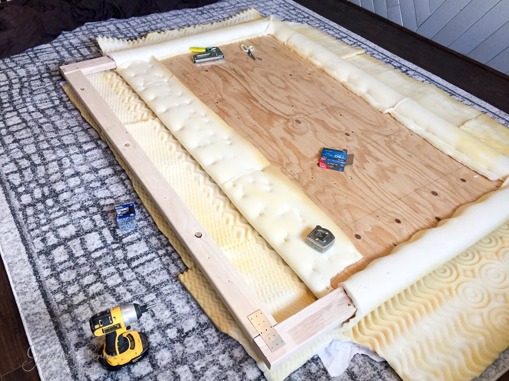 A DIY headboard project in progress, featuring a wooden frame with attached foam padding on a textured rug. A cordless drill and hardware packets are placed around the frame, with screws and brackets visible.