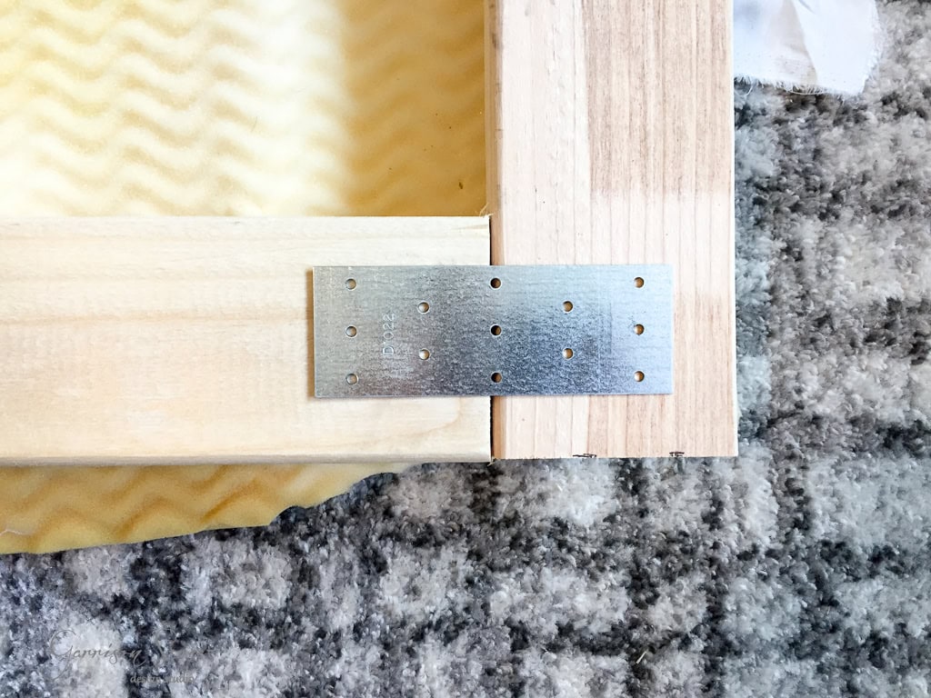 A close-up of a wooden frame corner secured with a metal bracket. The frame is placed on a gray and white textured carpet. The bracket has multiple holes for screws and the wood pieces are natural, light-colored timber.