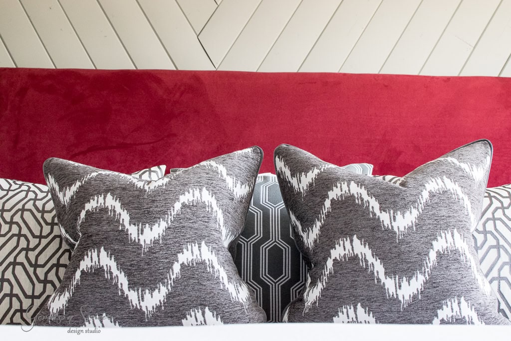 Two decorative pillows with a gray and white zigzag pattern are placed on a bed with a red headboard and a geometric patterned pillow behind them. The wall is designed with white wooden planks arranged diagonally.